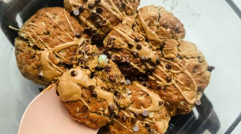 A glass dish containing seven chunky cookies drizzled with light brown icing and sprinkled with mini chocolate chips, one cookie being lifted with a pink spatula.