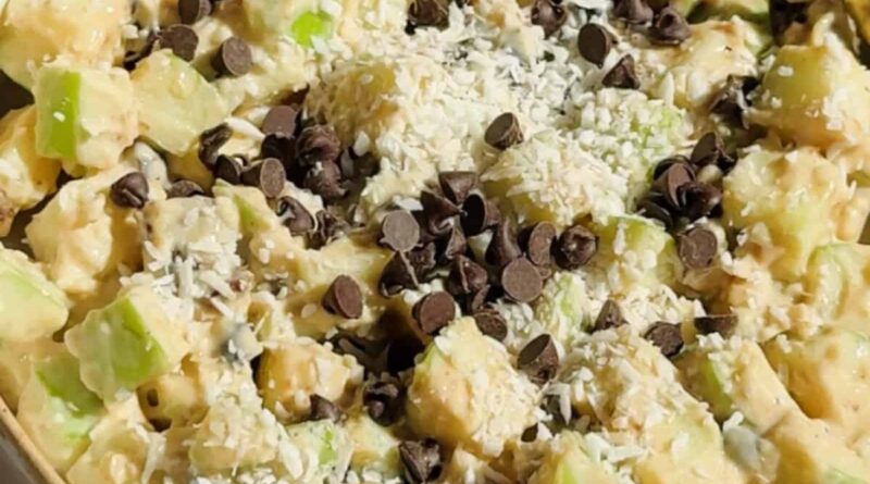 A bowl of chopped apples mixed with creamy sauce, topped with shredded coconut and chocolate chips, with a spoon on the side.