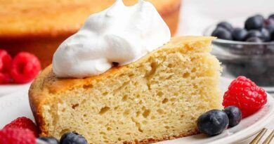 A slice of yellow cake topped with whipped cream, served on a plate with fresh raspberries and blueberries; whole cake and more berries in the background.