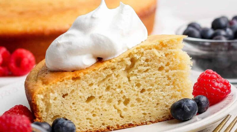 A slice of yellow cake topped with whipped cream, served on a plate with fresh raspberries and blueberries; whole cake and more berries in the background.