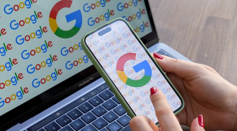 A woman looks at a mobile phone displaying the logo of Google in front of a laptop screen displaying the logo of Google.