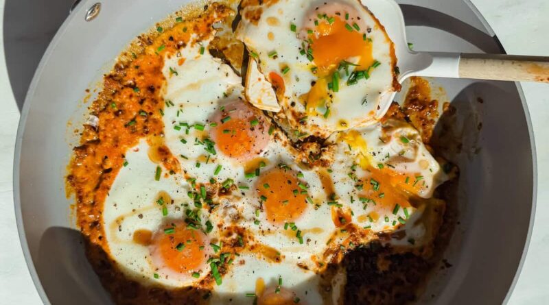 Fried eggs cooked in a skillet with tomato sauce and herbs, with a spatula lifting one egg.