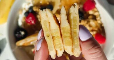 A hand with light purple nail polish holds three stacked pancake slices; a bowl with berries, granola, and syrup is blurred in the background.