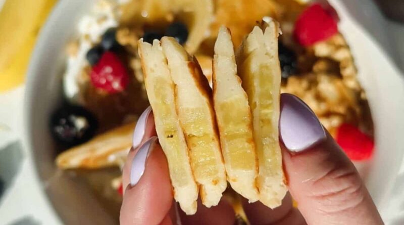A hand with light purple nail polish holds three stacked pancake slices; a bowl with berries, granola, and syrup is blurred in the background.