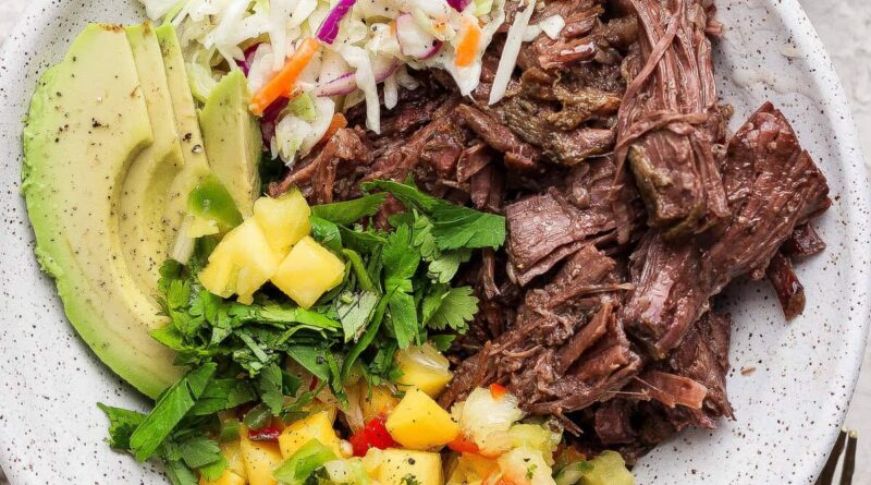 A bowl containing shredded beef, sliced avocado, coleslaw, chopped cilantro, and pineapple salsa.