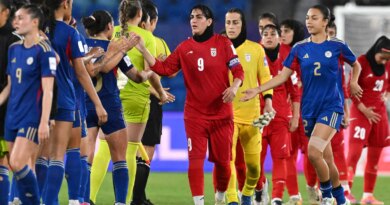 People in mostly ed and blue uniforms greet each other in two lines on a green field. A person in a red uniform with “No. 9” is in the center foreground.