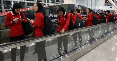 Women wearing red shirts and black head coverings stand on a moving walkway, some looking at phones.