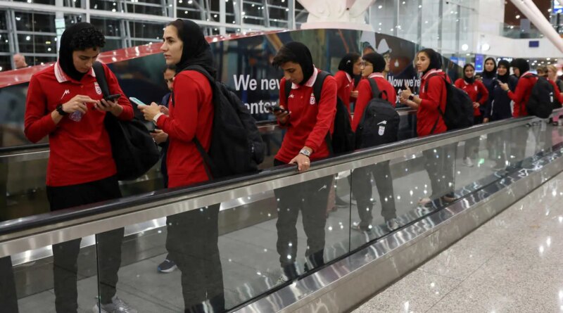 Women wearing red shirts and black head coverings stand on a moving walkway, some looking at phones.