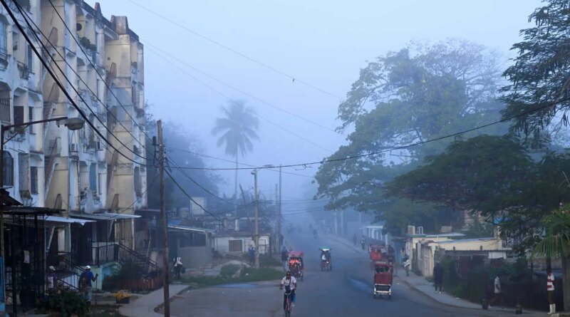 A hazy street scene shows a curved road lined with buildings and large trees. People move on foot, bicycle, and in three-wheeled vehicles, as a rooster walks in the foreground.