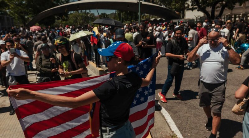 A person in a red-and-blue cap holds a large American flag. A crowd fills the background.