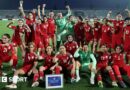 Players from the Afghan Women United team, half standing and half seated, pose for a post-match photo, smiling and with fists raised
