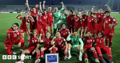 Players from the Afghan Women United team, half standing and half seated, pose for a post-match photo, smiling and with fists raised