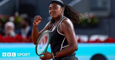 Hailey Baptiste, wearing a black vest and shorts, black headband and a ponytail, holds her green Wilson racket in her left hand and punches her right fist in celebration