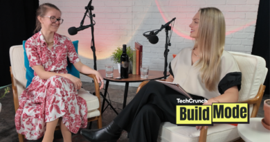 Two women sit facing each other in a podcast studio, speaking into microphones. One wears a red floral dress and glasses, while the other wears a cream top and black pants. A small table between them holds a bottle of wine, glasses of water, and books. A TechCrunch Build Mode logo appears in the corner.