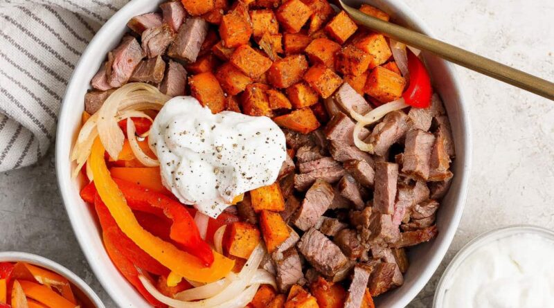 A bowl filled with diced steak, roasted sweet potatoes, saut&eacute;ed onions and bell peppers, topped with a dollop of sour cream and black pepper, with a fork resting inside.