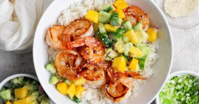 A bowl of white rice topped with grilled shrimp, diced mango, cucumber, avocado, and sprinkled sesame seeds, with side bowls of salsa and chopped green onions.