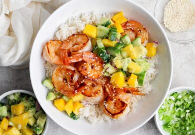 A bowl of white rice topped with grilled shrimp, diced mango, cucumber, avocado, and sprinkled sesame seeds, with side bowls of salsa and chopped green onions.