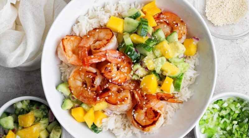 A bowl of white rice topped with grilled shrimp, diced mango, cucumber, avocado, and sprinkled sesame seeds, with side bowls of salsa and chopped green onions.