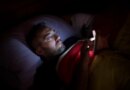 A man in glasses lies in bed, illuminated by the soft glow of his smartphone screen.