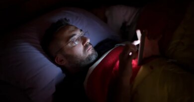 A man in glasses lies in bed, illuminated by the soft glow of his smartphone screen.