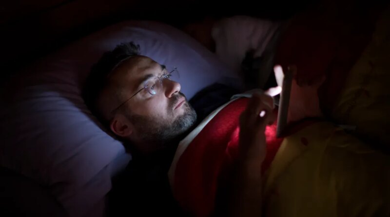 A man in glasses lies in bed, illuminated by the soft glow of his smartphone screen.