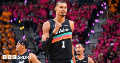 Victor Wembanyama, wearing a black San Antonio vest with a turquoise, pink, and orange stripe across the chest, punches his right fist in celebration and smiles