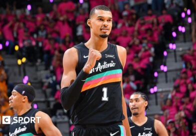 Victor Wembanyama, wearing a black San Antonio vest with a turquoise, pink, and orange stripe across the chest, punches his right fist in celebration and smiles