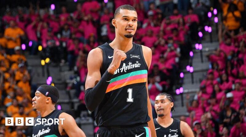 Victor Wembanyama, wearing a black San Antonio vest with a turquoise, pink, and orange stripe across the chest, punches his right fist in celebration and smiles