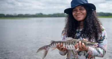 A young woman wearing a broad hat holds a fish next to a river, smiling 
