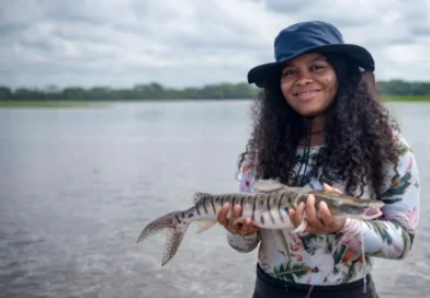 A young woman wearing a broad hat holds a fish next to a river, smiling 