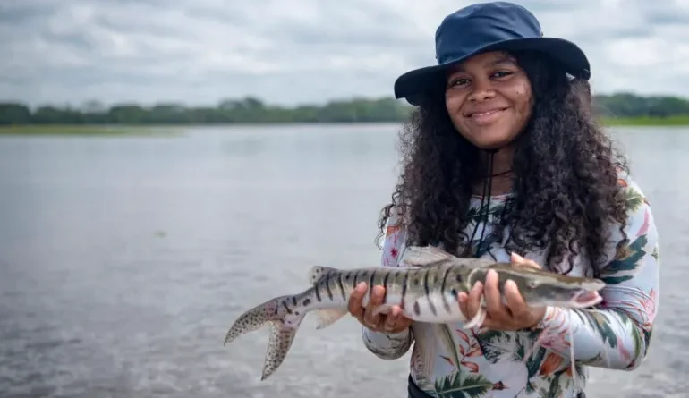 A young woman wearing a broad hat holds a fish next to a river, smiling