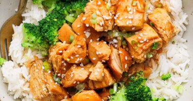 A bowl of white rice topped with cooked chicken pieces in sauce, broccoli florets, and a sprinkle of sesame seeds, with a fork on the side.