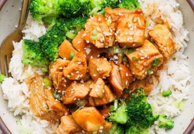 A bowl of white rice topped with cooked chicken pieces in sauce, broccoli florets, and a sprinkle of sesame seeds, with a fork on the side.