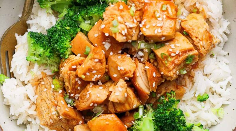 A bowl of white rice topped with cooked chicken pieces in sauce, broccoli florets, and a sprinkle of sesame seeds, with a fork on the side.