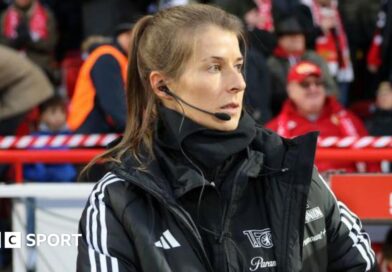 Marie-Louise Eta, wearing a black coat and top with a black microphone and headset in her right ear, on the touchline with fans in red and dark clothing in the background