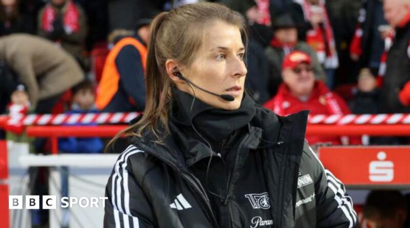 Marie-Louise Eta, wearing a black coat and top with a black microphone and headset in her right ear, on the touchline with fans in red and dark clothing in the background
