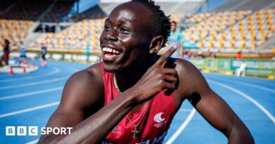 Gout Gout, wearing a red vest and a gold crucifix around his neck, points with his right hand across his body and smiles while crouching on a blue athletics track with white and yellow stadium seating in the background.