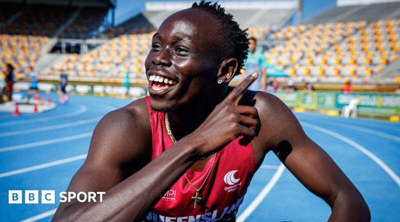 Gout Gout, wearing a red vest and a gold crucifix around his neck, points with his right hand across his body and smiles while crouching on a blue athletics track with white and yellow stadium seating in the background.