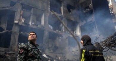 Two emergency workers look up at a damaged building with smoke. One wears a camouflage uniform; the other, a dark jacket with yellow stripes and text.