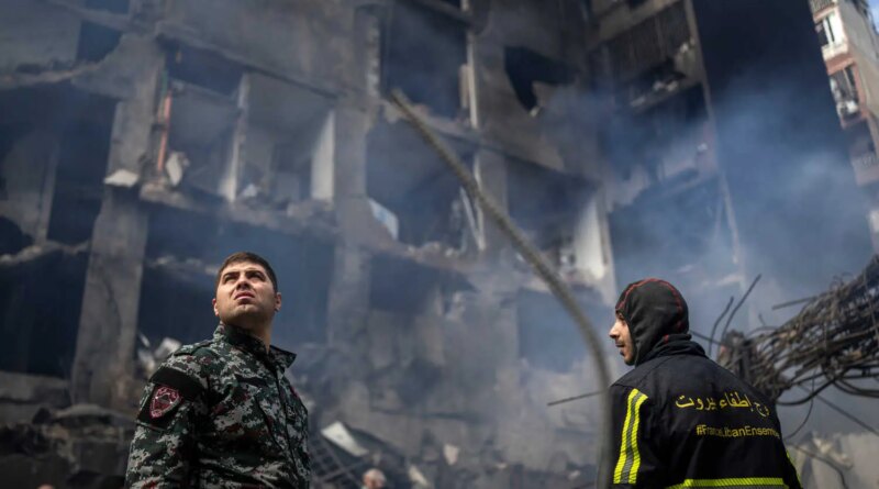 Two emergency workers look up at a damaged building with smoke. One wears a camouflage uniform; the other, a dark jacket with yellow stripes and text.