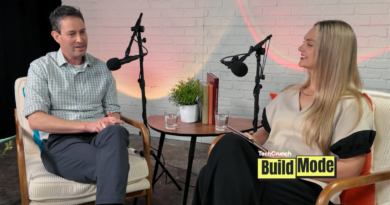 A podcast-style interview setup shows a man and a woman seated across from each other in armchairs, speaking into microphones. They sit beside a small round table with water glasses, books, and a potted plant, against a white brick wall backdrop. A “TechCrunch Build Mode” logo appears in the lower corner.