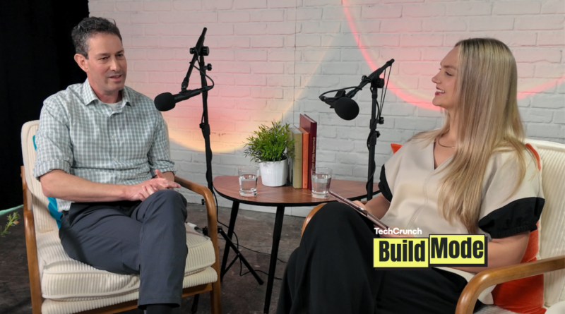 A podcast-style interview setup shows a man and a woman seated across from each other in armchairs, speaking into microphones. They sit beside a small round table with water glasses, books, and a potted plant, against a white brick wall backdrop. A “TechCrunch Build Mode” logo appears in the lower corner.