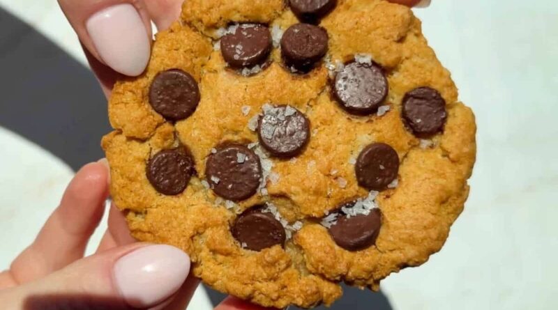 A close-up of hands holding a chocolate chip cookie with visible sea salt flakes on top.