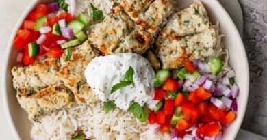 A bowl of white rice topped with grilled kebab pieces, diced red onion, red and green bell peppers, and a dollop of yogurt garnished with a mint leaf.