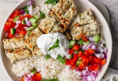 A bowl of white rice topped with grilled kebab pieces, diced red onion, red and green bell peppers, and a dollop of yogurt garnished with a mint leaf.