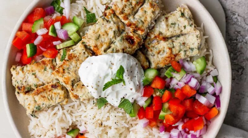 A bowl of white rice topped with grilled kebab pieces, diced red onion, red and green bell peppers, and a dollop of yogurt garnished with a mint leaf.