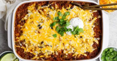 A baked casserole topped with melted cheese, black beans, sour cream, and chopped cilantro, surrounded by bowls of lime, shredded cheese, and extra sour cream.