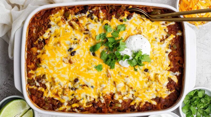 A baked casserole topped with melted cheese, black beans, sour cream, and chopped cilantro, surrounded by bowls of lime, shredded cheese, and extra sour cream.