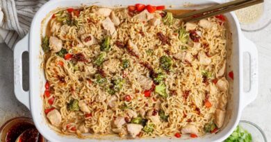 Rectangular white baking dish filled with cooked ramen noodles, chicken pieces, broccoli, and red peppers, garnished with sesame seeds and sauce, with chopsticks resting on the dish.