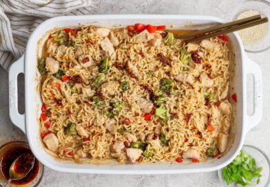 Rectangular white baking dish filled with cooked ramen noodles, chicken pieces, broccoli, and red peppers, garnished with sesame seeds and sauce, with chopsticks resting on the dish.